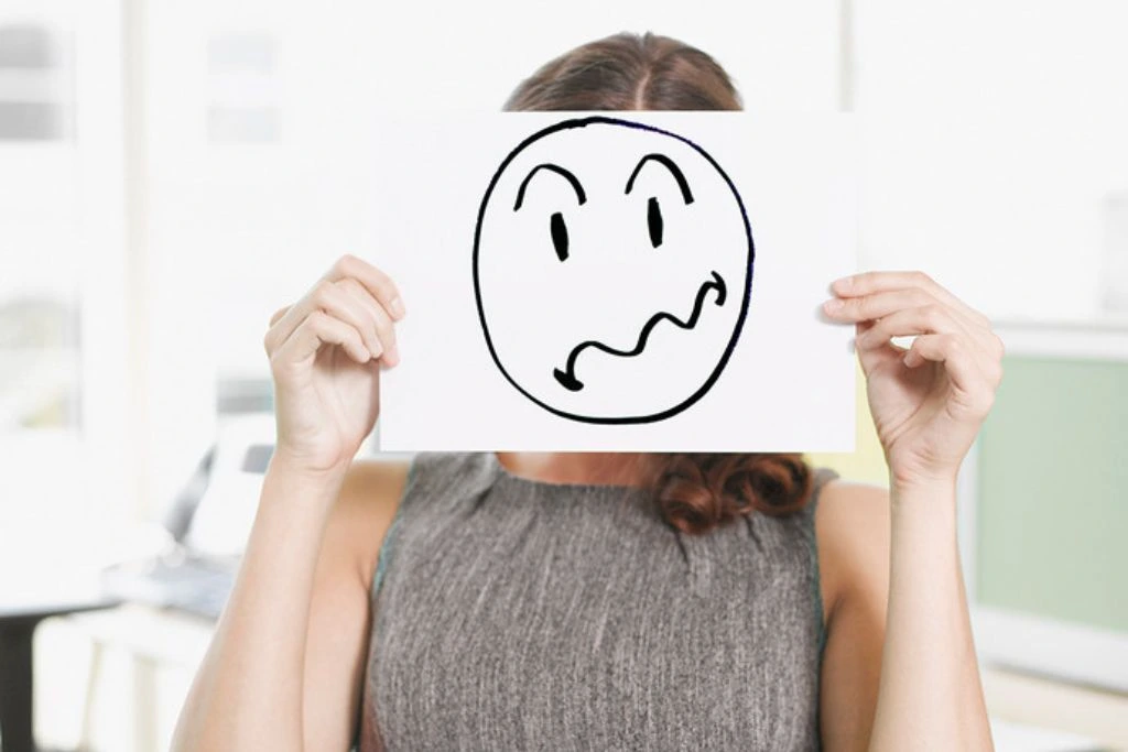A woman holding a paper with a drawing of a disgruntled emoji that covers her face.