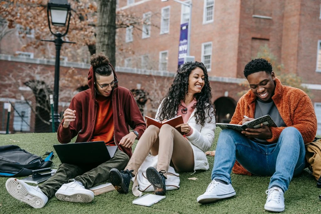 Three college students compare notes while sitting on a field just outside the university.