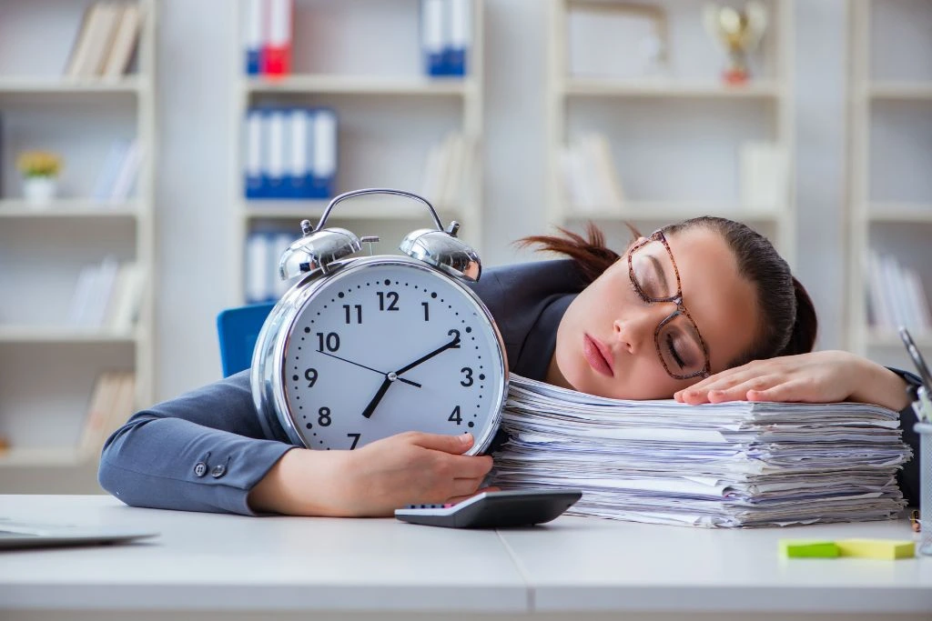 A woman dozing off on top of a pile of papers while holding a huge alarm clock.