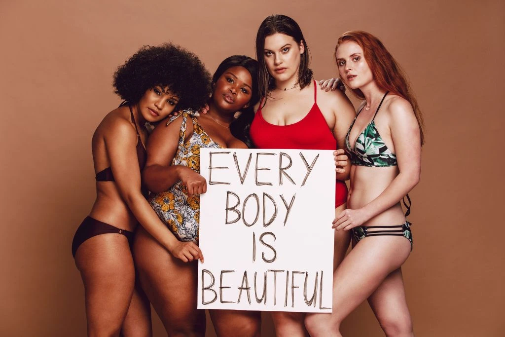 Four women with a different body type holding a sign that says "Every body is beautiful"