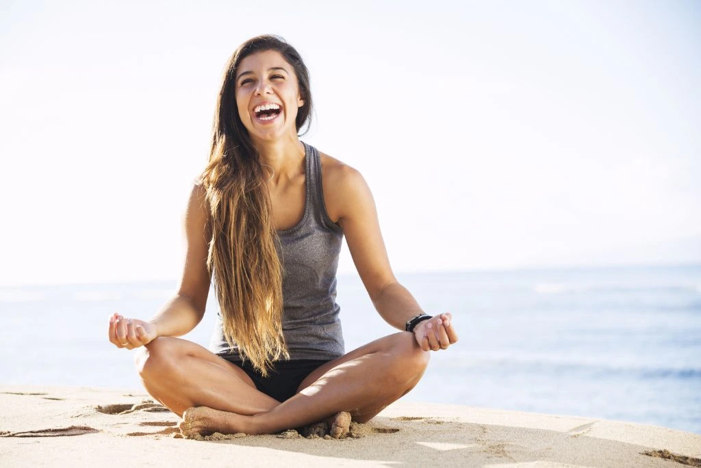 A woman doing the meditation position having a laugh by the beach.