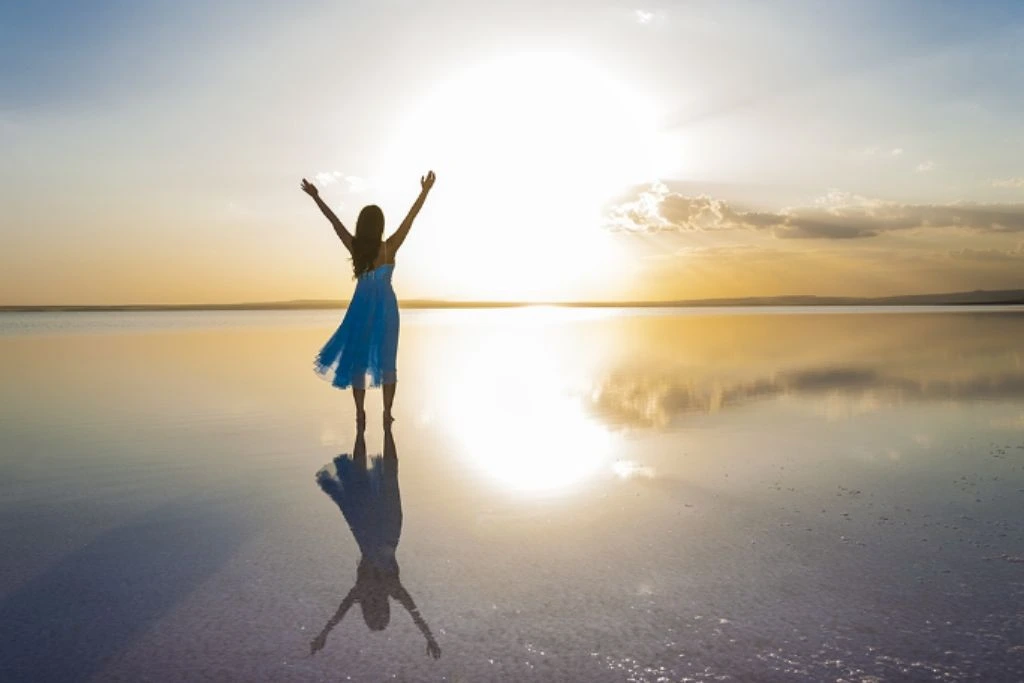 A woman standing on top of a shallow salt lake that cast a perfect reflection of her raising her hands facing the sun sets.