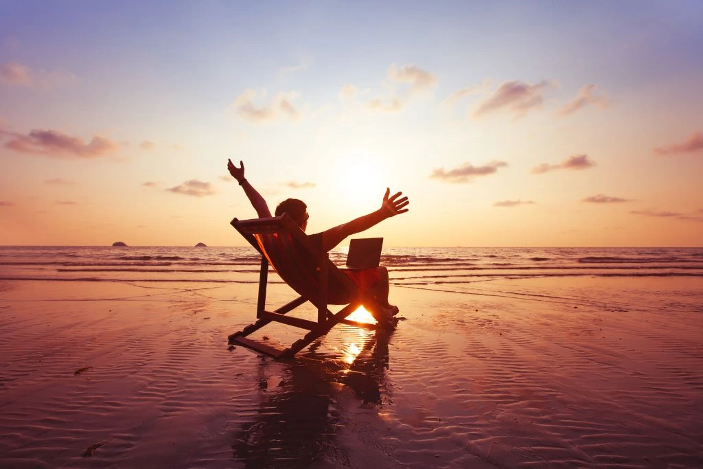 A man sitting with a laptop on his lap, in front of the ocean while his arm are wide open that seems to hug the sunset.