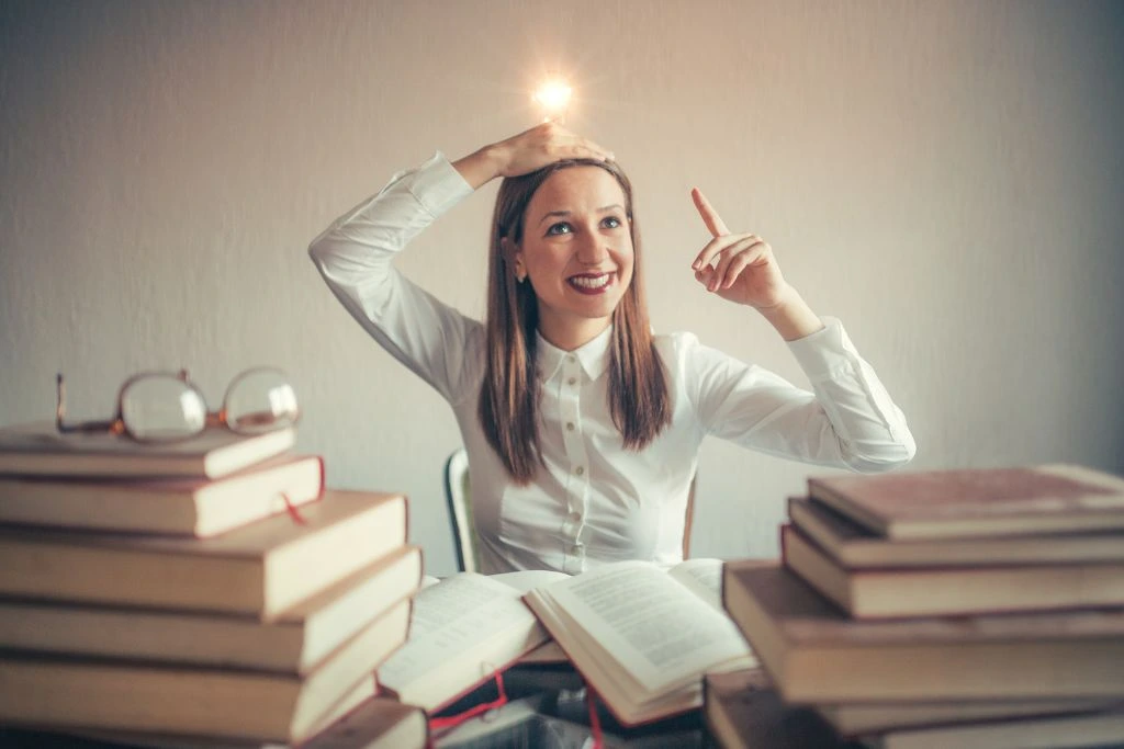 A women that appears to have a light bulb above her head while sitting in front of piled books