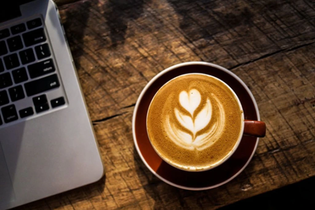A nice cup of coffee with a heart and leaf shape of its froth beside a laptop.