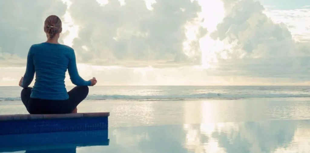 An image of a woman sitting on a mantra post in front of an infinity pool facing the beach.
