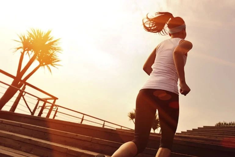 A woman dressed in an athletic outfit is running up a long stair under the heat of the sun.